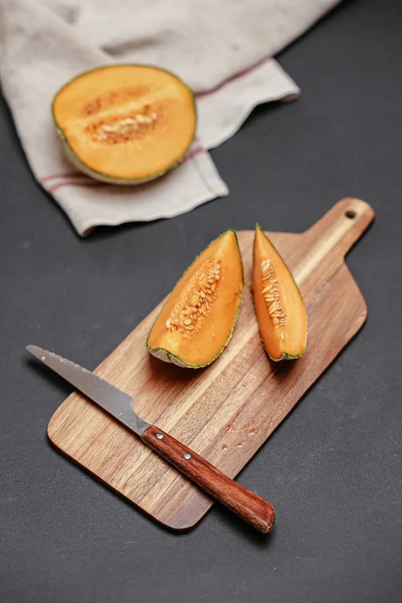 Rodajas de melón fresco cortado en cubos sobre una tabla de madera junto a hojas verdes y luz natural